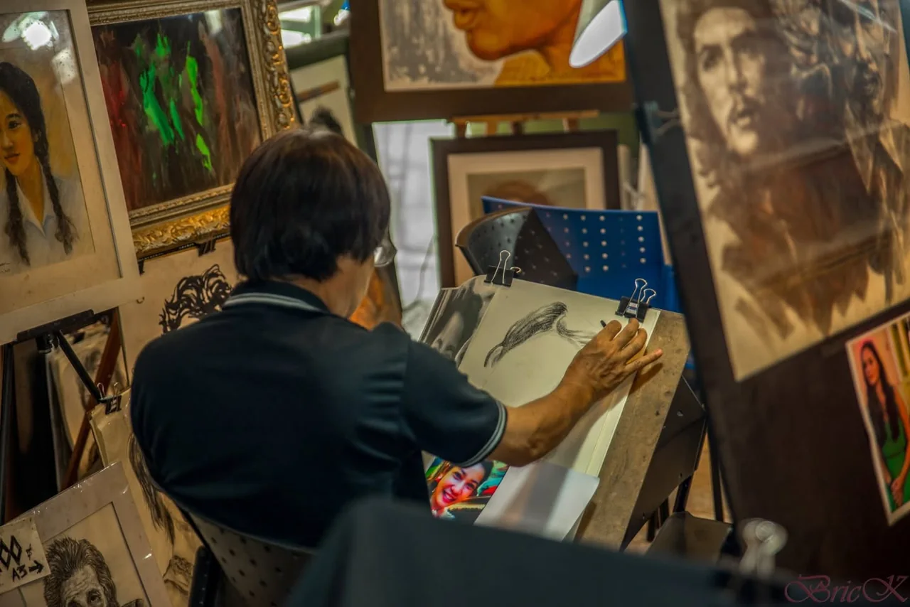 An artist draws at an easel surrounded by portraits in a workshop.