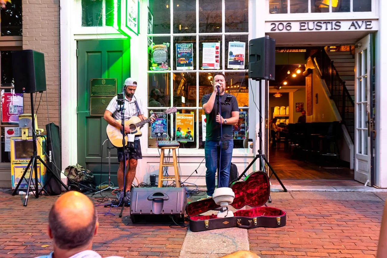 Two musicians perform on a sidewalk in front of a colorful venue.