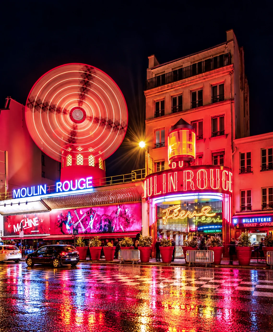 Bright red Moulin Rouge lights reflect on a wet city street at night.