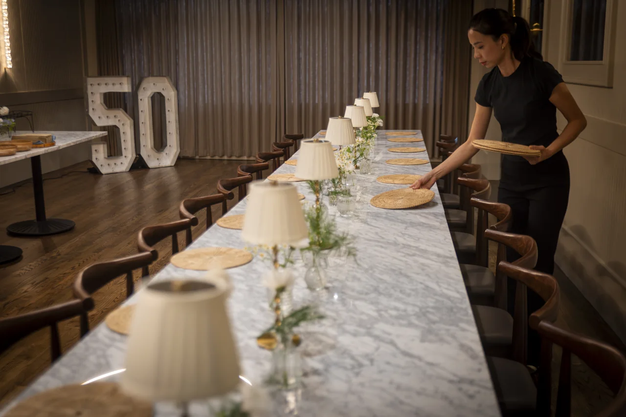 A member of staff arranges a long dining table with flowers and lamps.