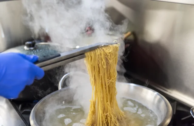 Steaming noodles are lifted above a pan in a busy kitchen.
