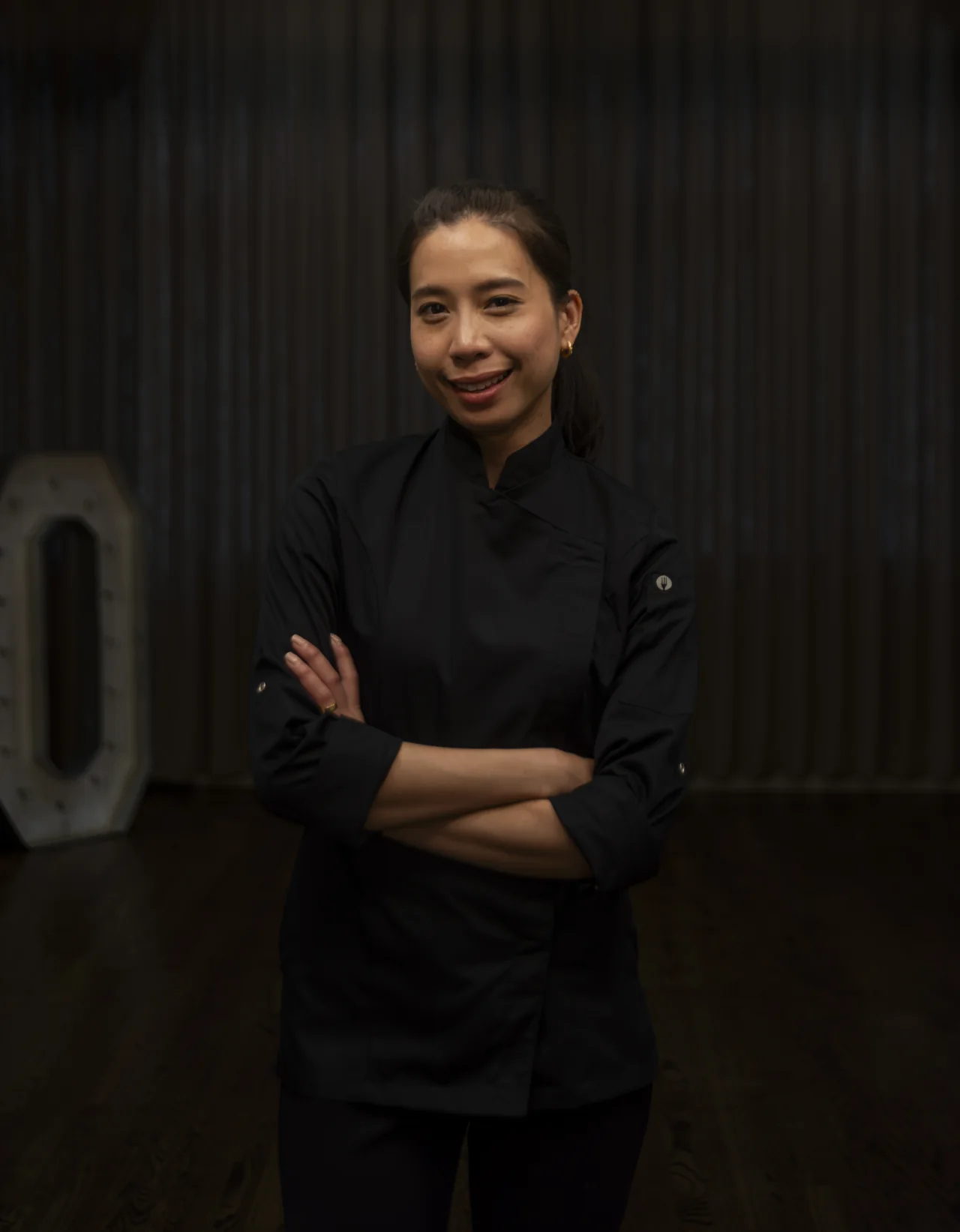A chef stands with folded arms in a warmly lit restaurant kitchen.