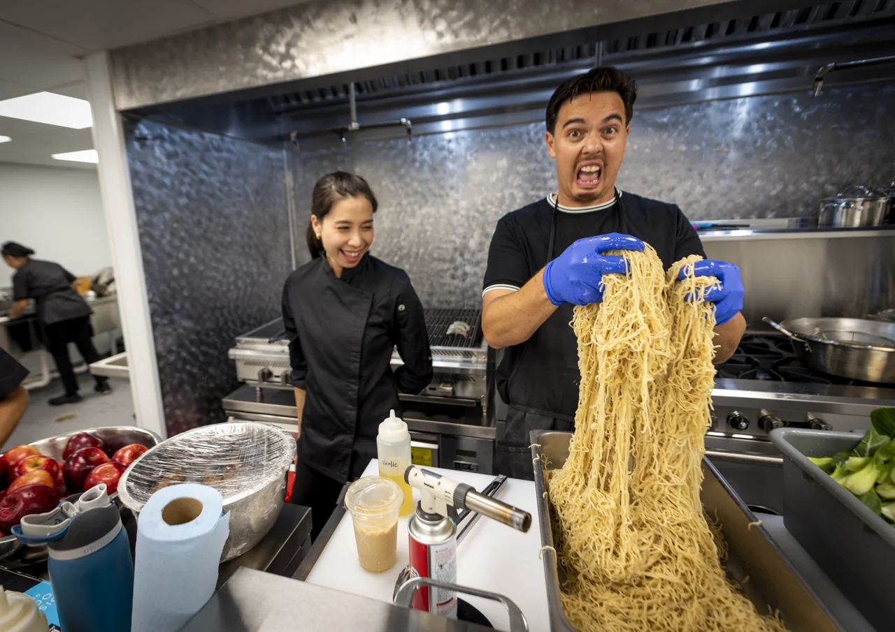 A smiling chef lifts fresh noodles while working in a kitchen.