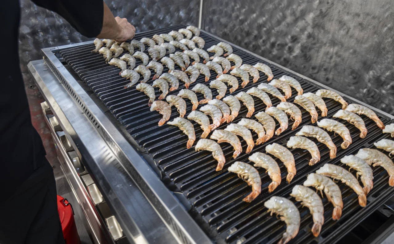 Rows of shrimp cook together on a dark grill.