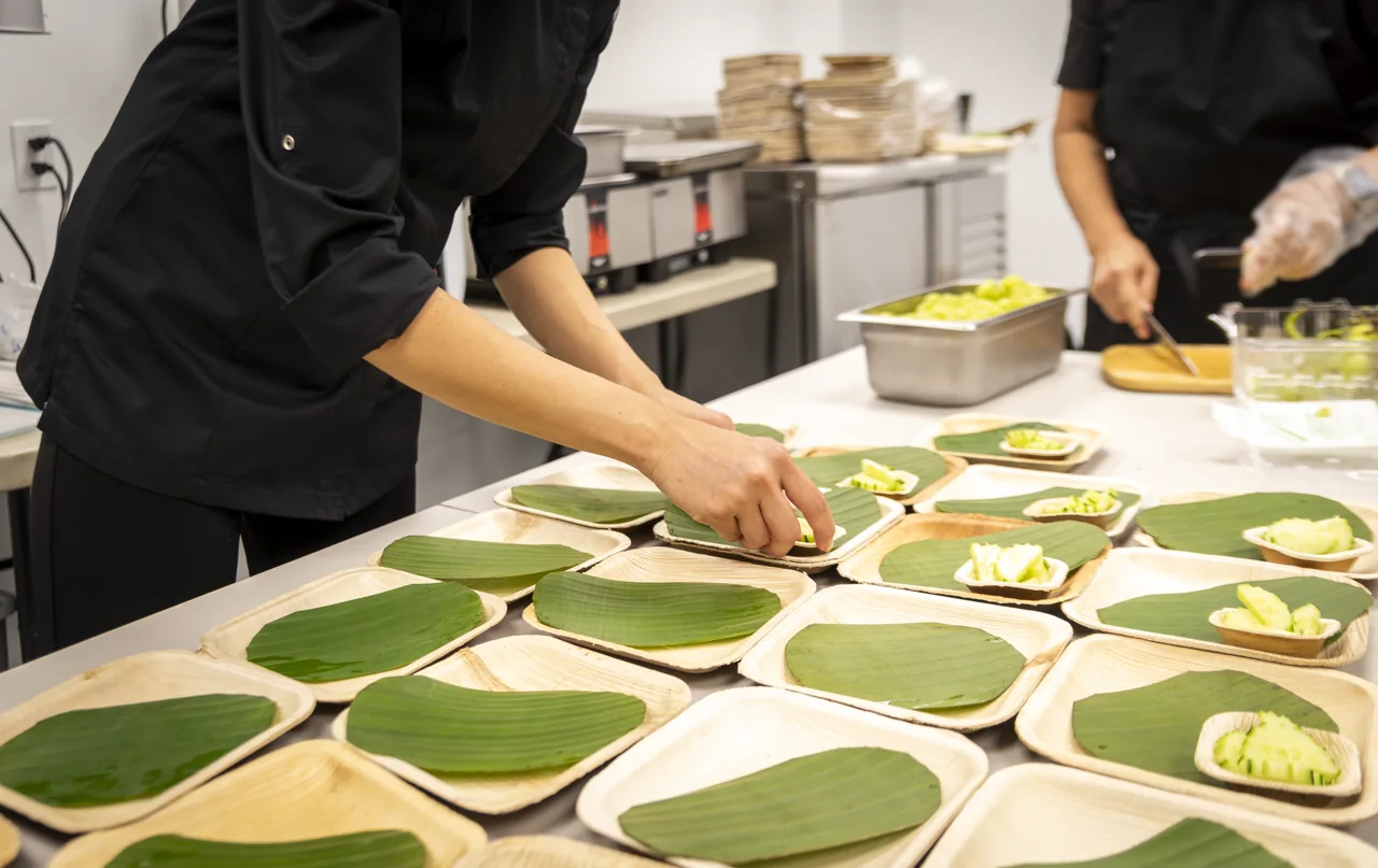 Hands arrange green leaves across rows of plates in a kitchen.