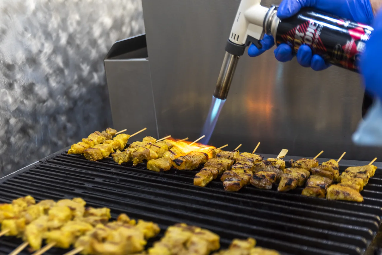 A cook uses a blue flame to finish rows of food on a grill.