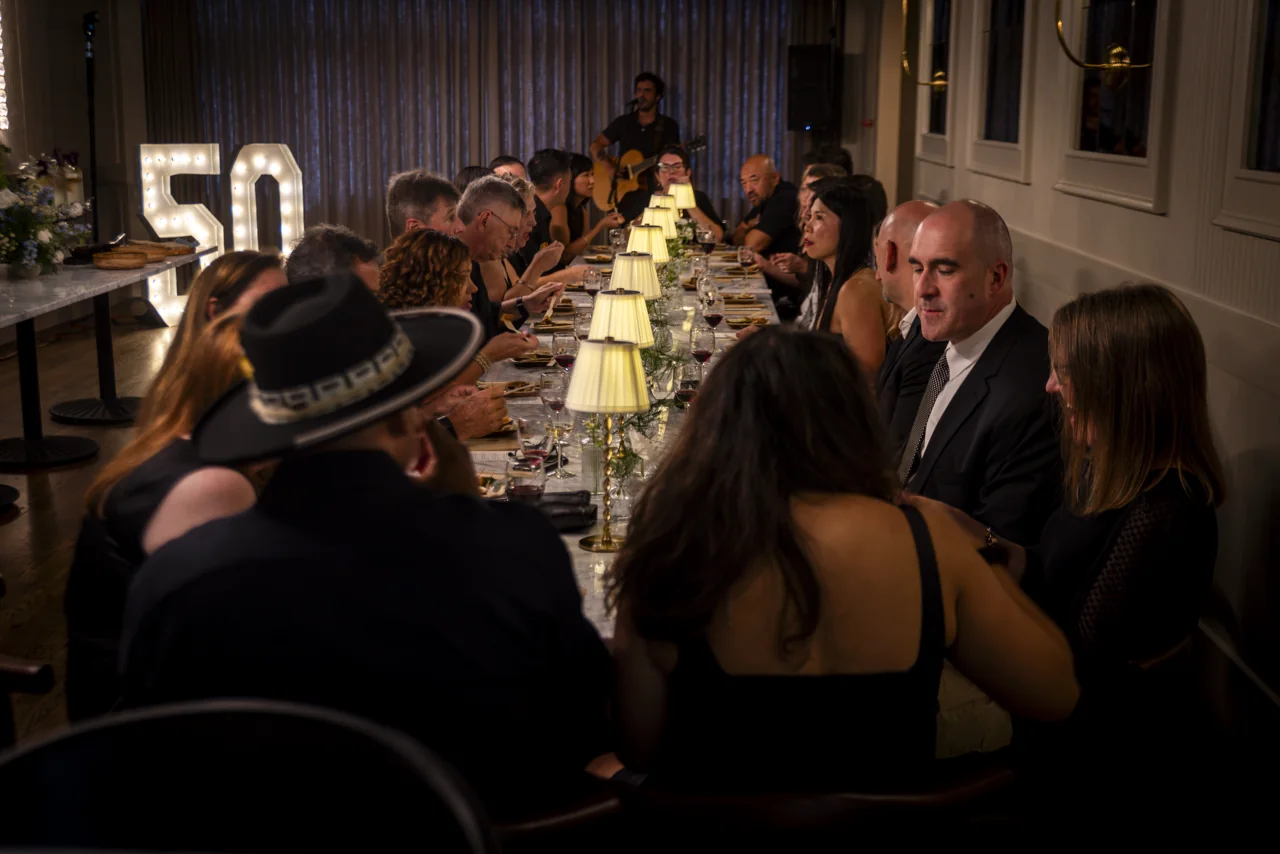Guests share a long restaurant table lit by small warm lamps.