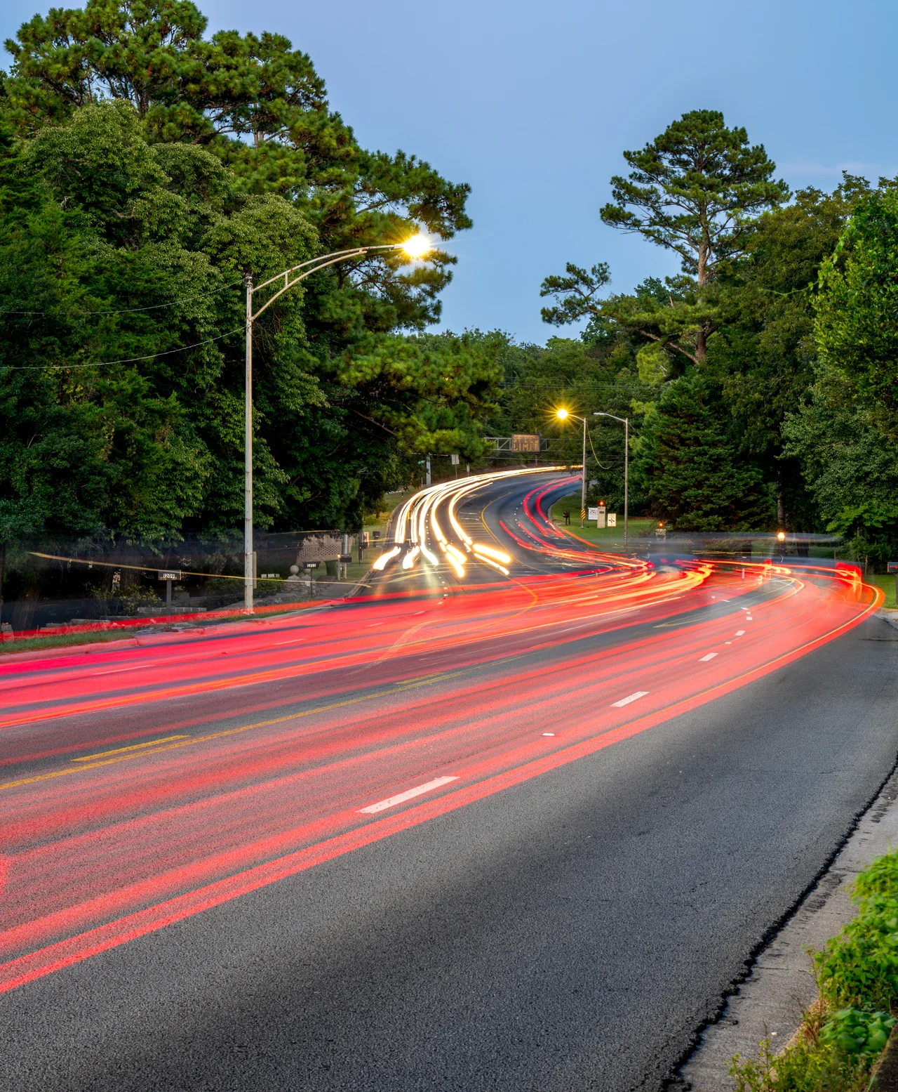 Red light trails sweep around a tree-lined road beneath a pale evening sky.