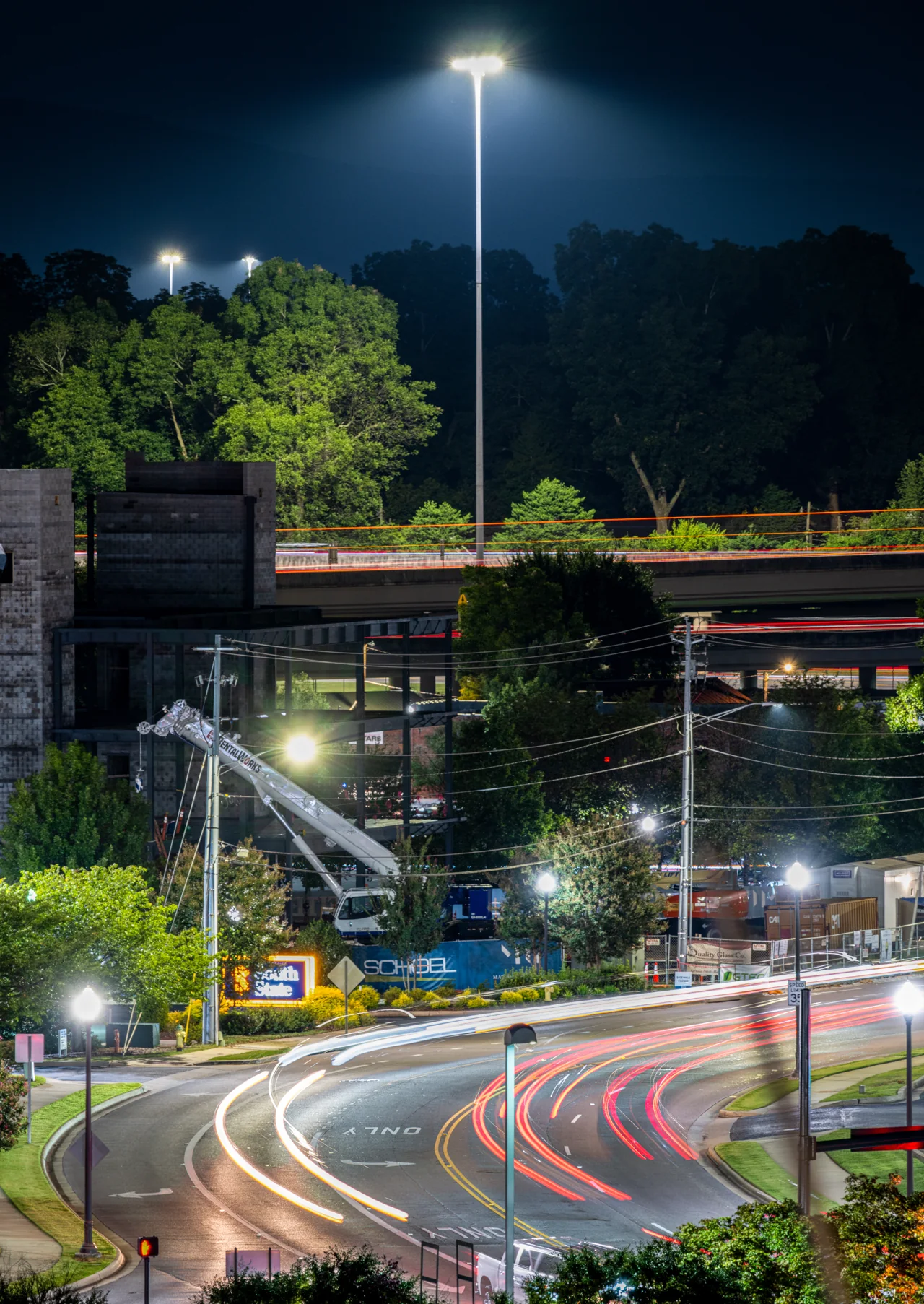 Red and white traffic trails curve beside water and trees beneath a bright streetlight.