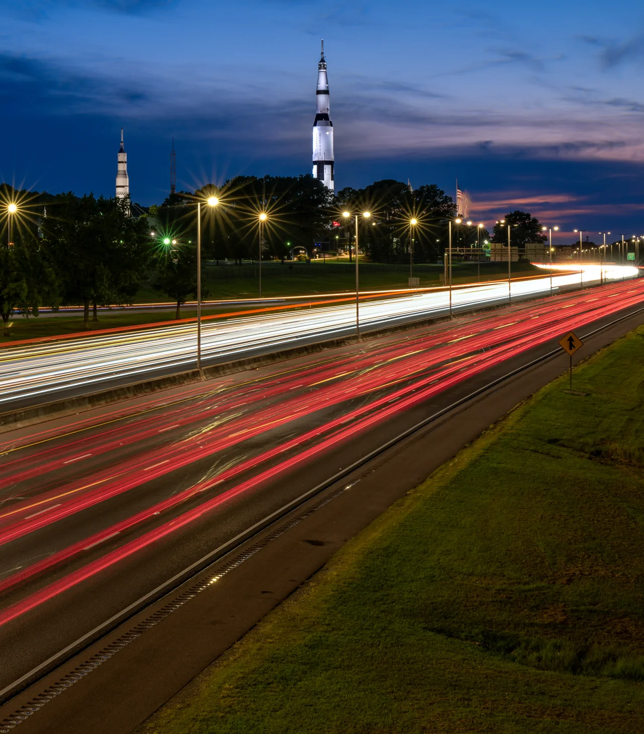 Red and white traffic trails cross a road beneath a rocket-shaped landmark at dusk.