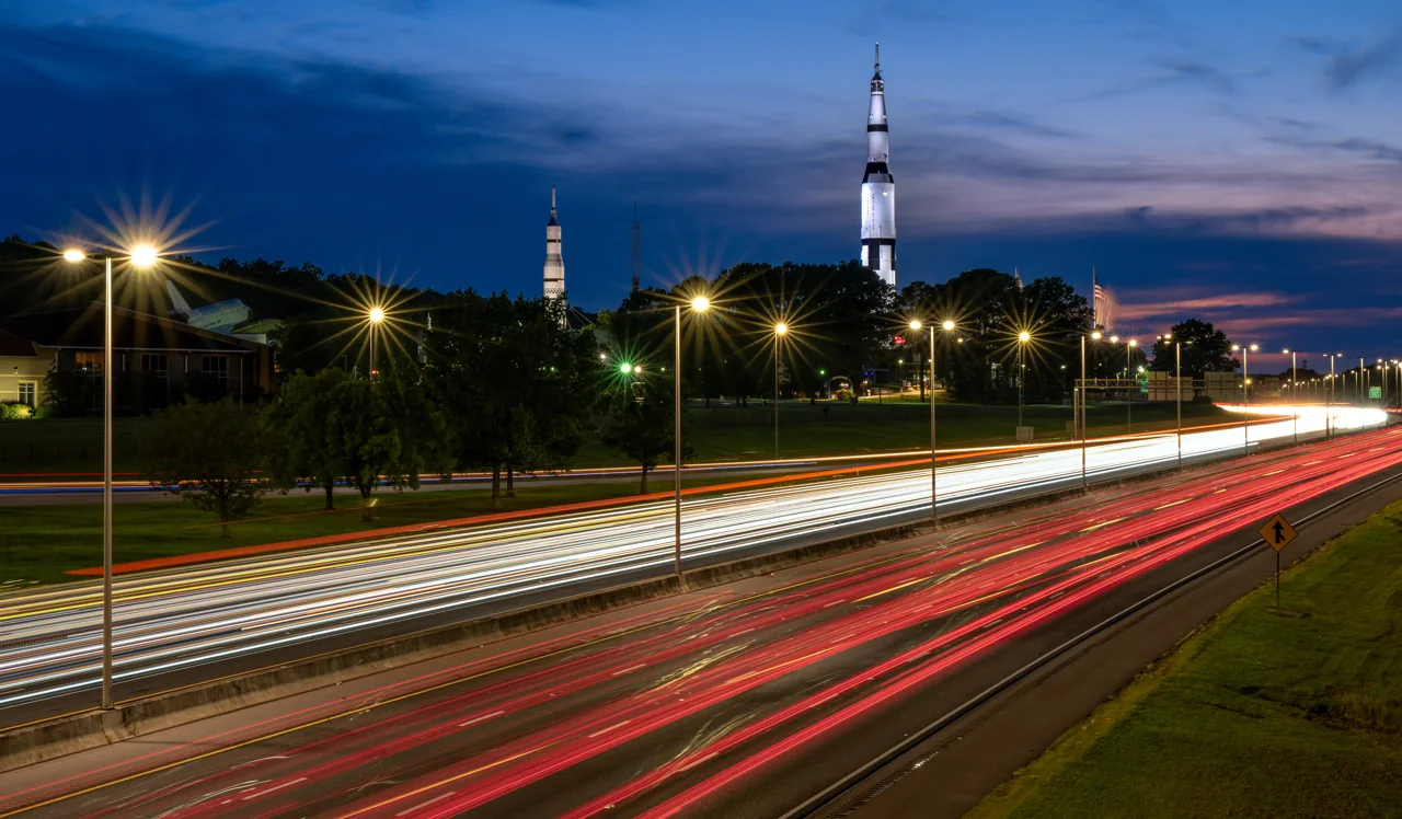 A wide view of red and white traffic trails beneath a blue evening sky and illuminated rocket-shaped landmark.