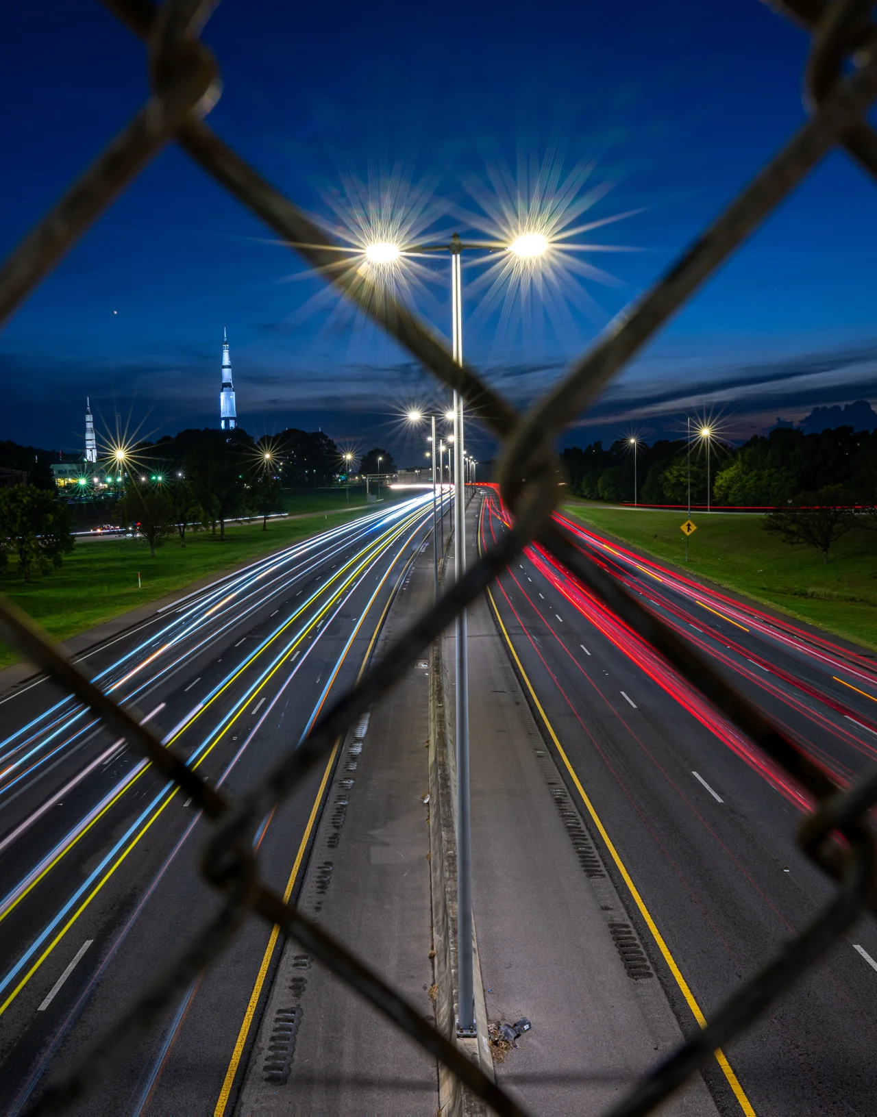 Night traffic trails are seen through diagonal chain-link fencing.