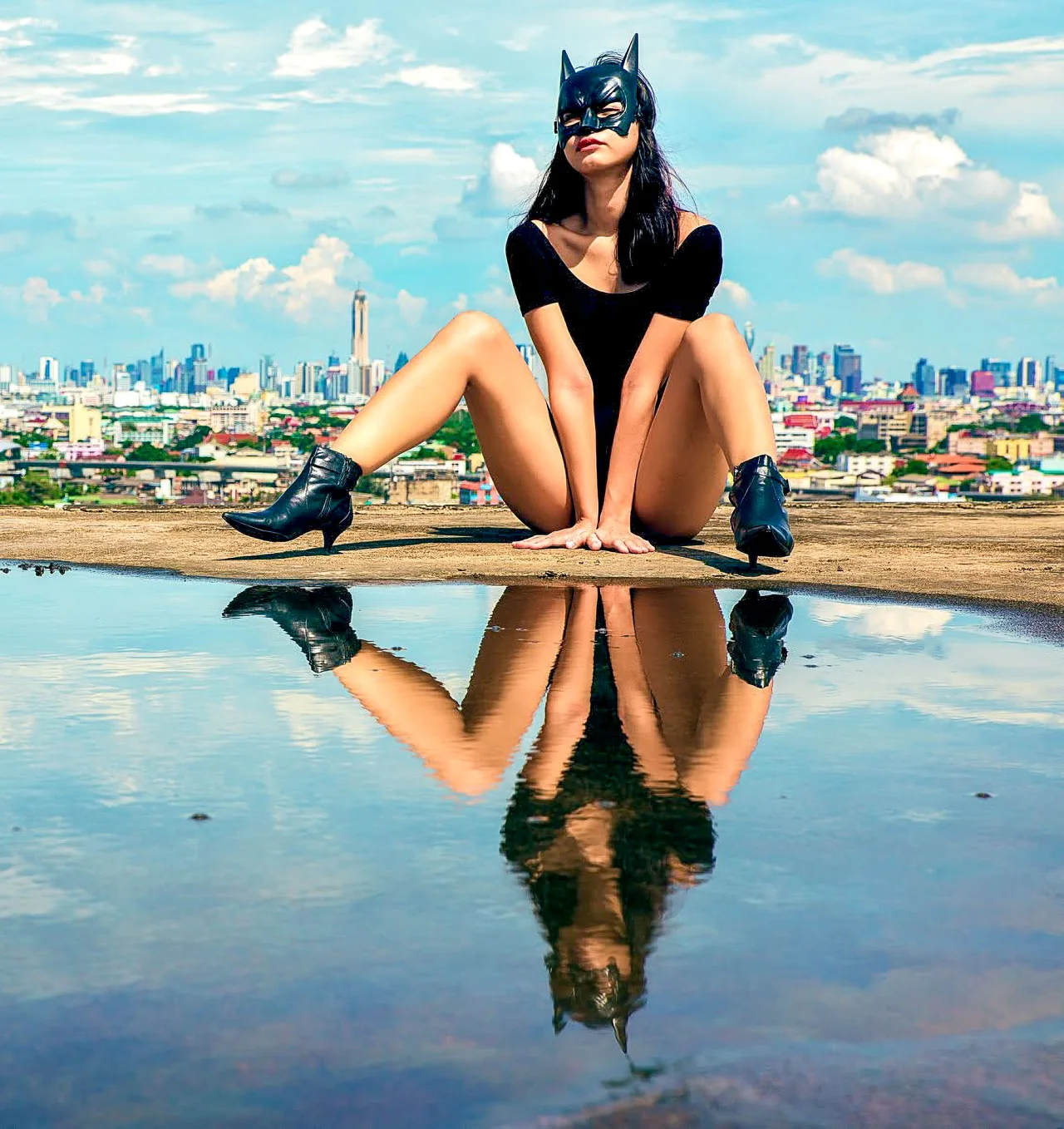 A seated woman in black wears a dark mask in a rooftop reflection.