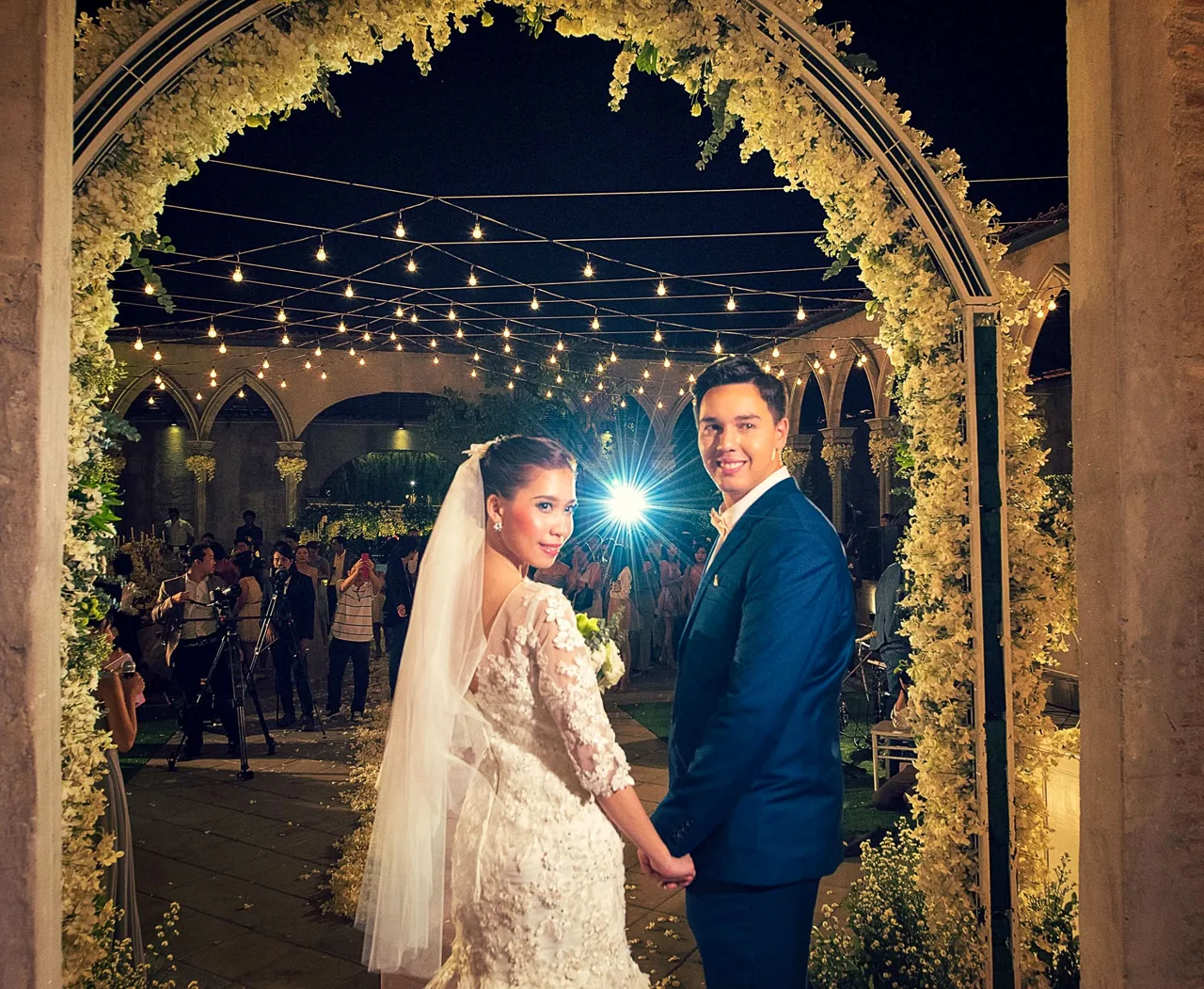 A bride and groom stand together beneath an outdoor arch.