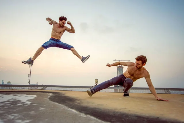A martial artist leaps toward another performer on an urban rooftop.