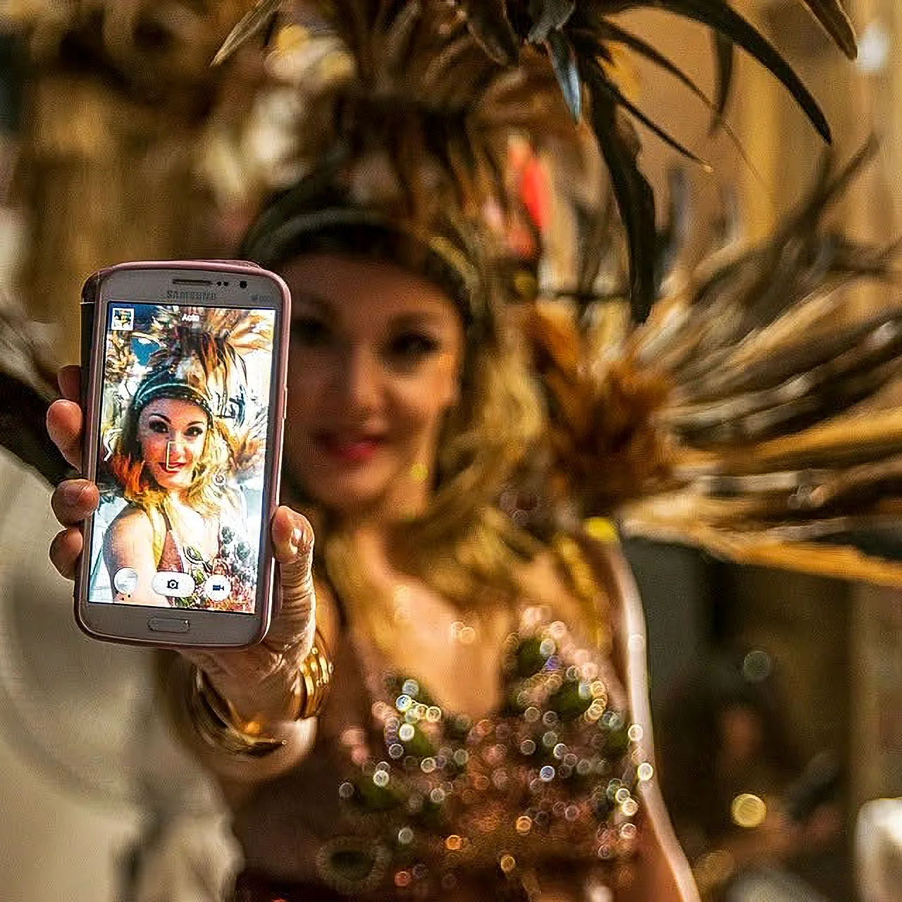 A woman in an elaborate feather costume holds a phone displaying a portrait.