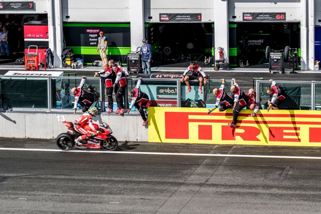 A motorcycle rider performs a wheelie beside a pit lane.