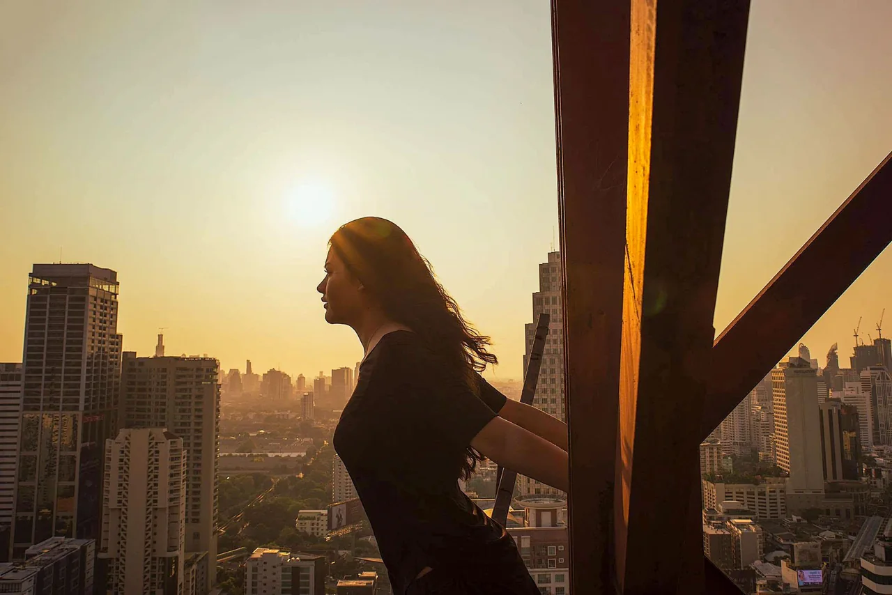 A woman in profile leans beside an opening overlooking a city in golden evening light.