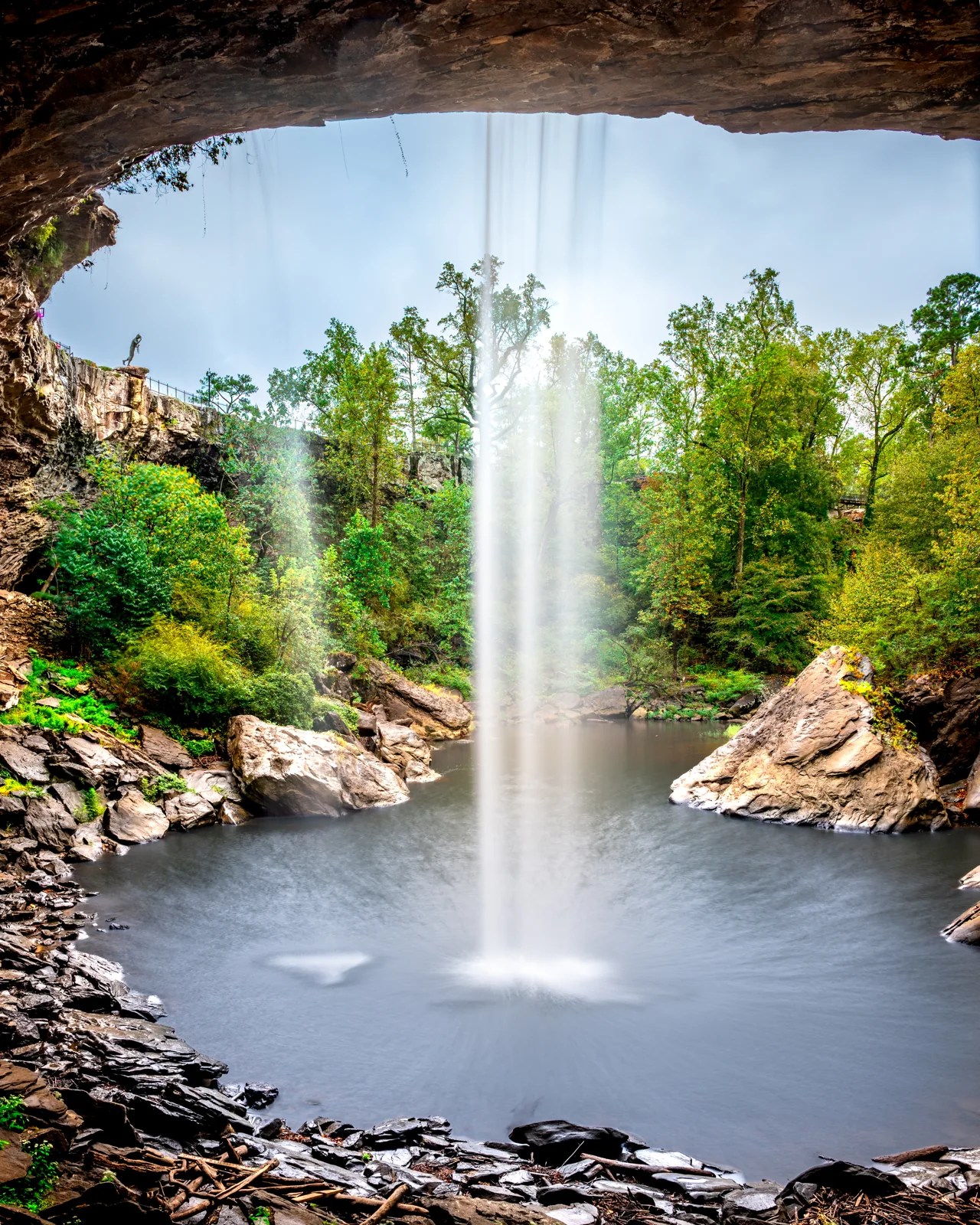 A waterfall and woodland are framed by a dark rock overhang.