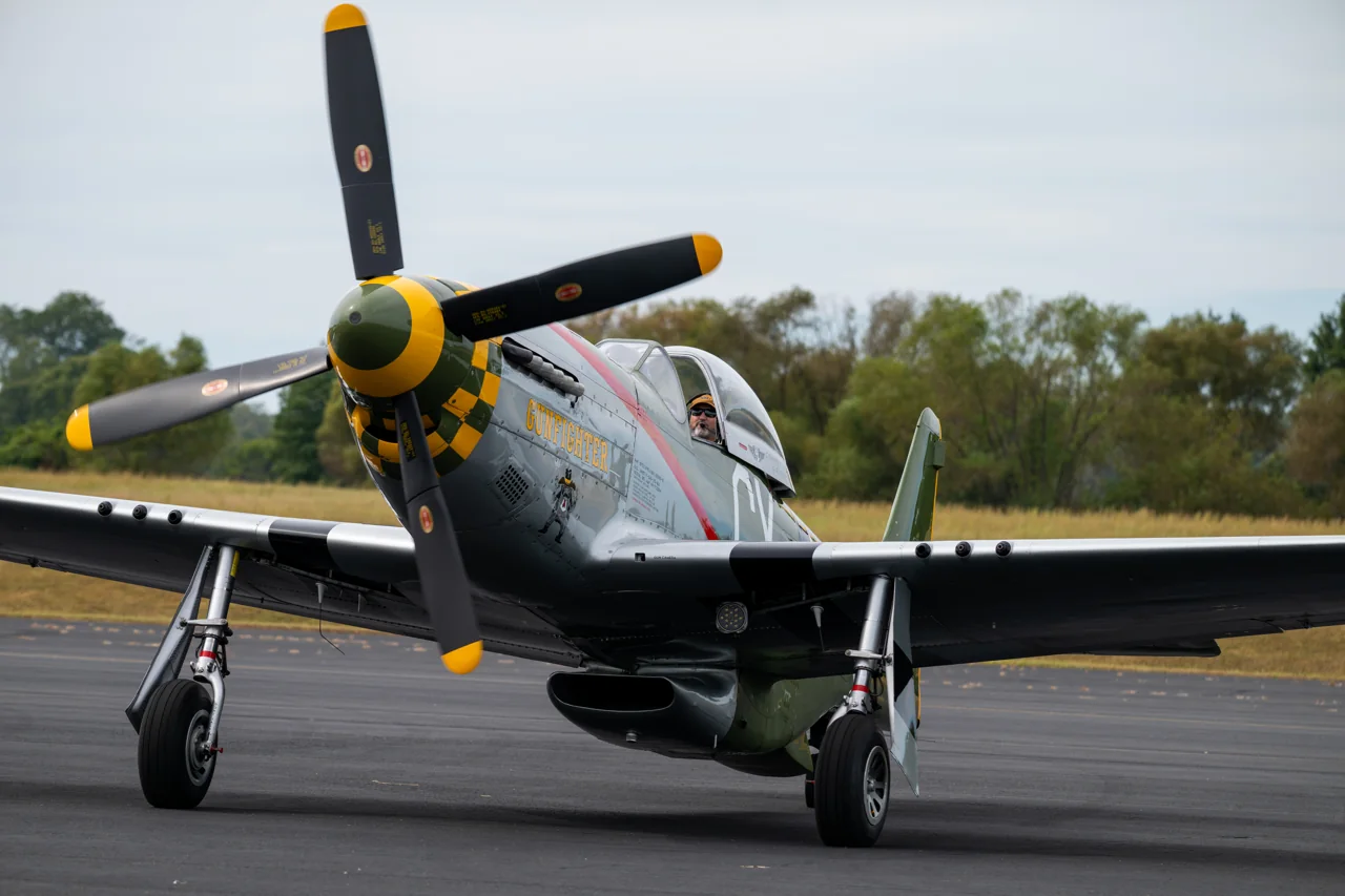 A vintage aircraft faces the camera with its propeller and shadow across the runway.